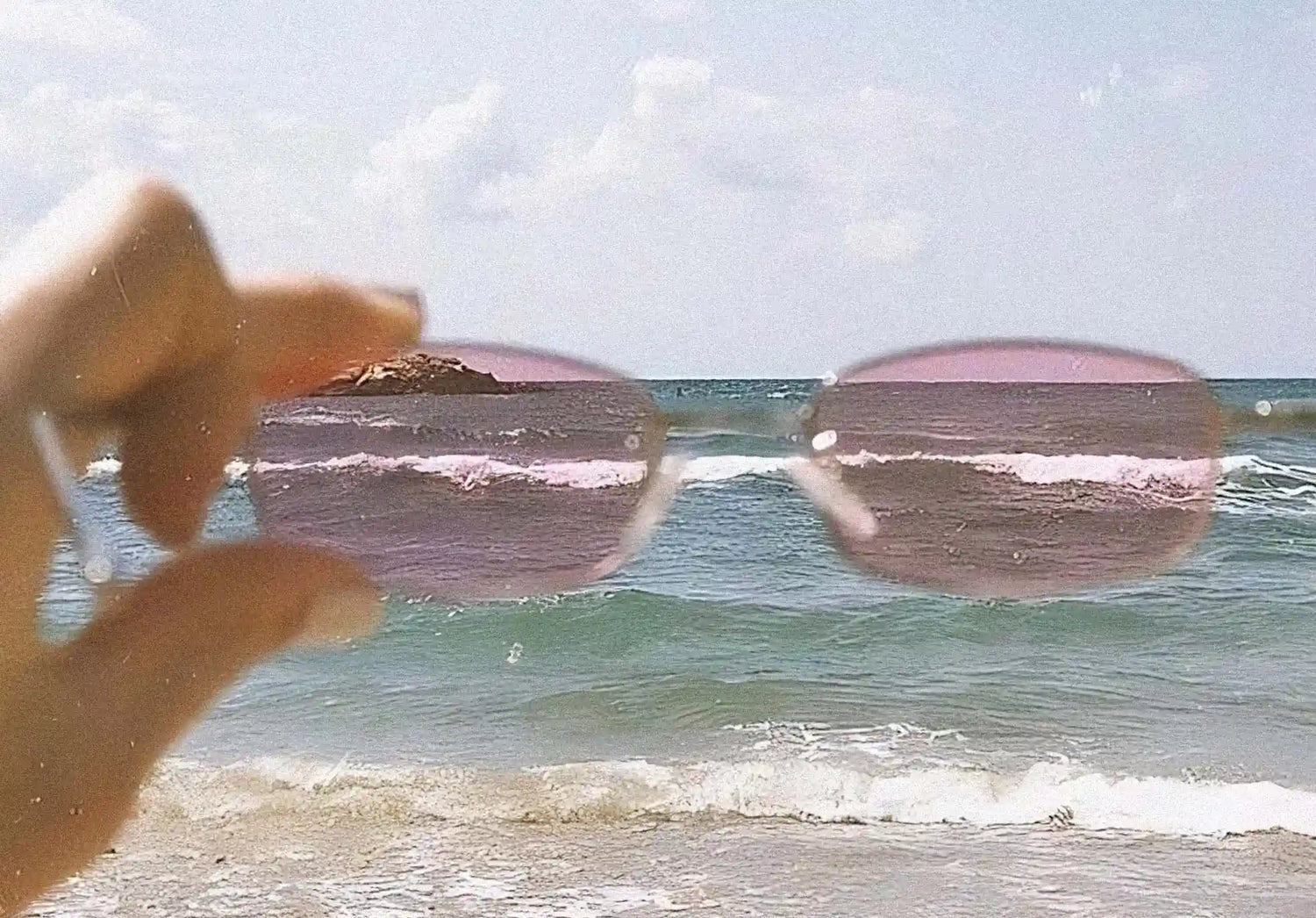 View of the beach and ocean through rose-colored glasses held by a hand